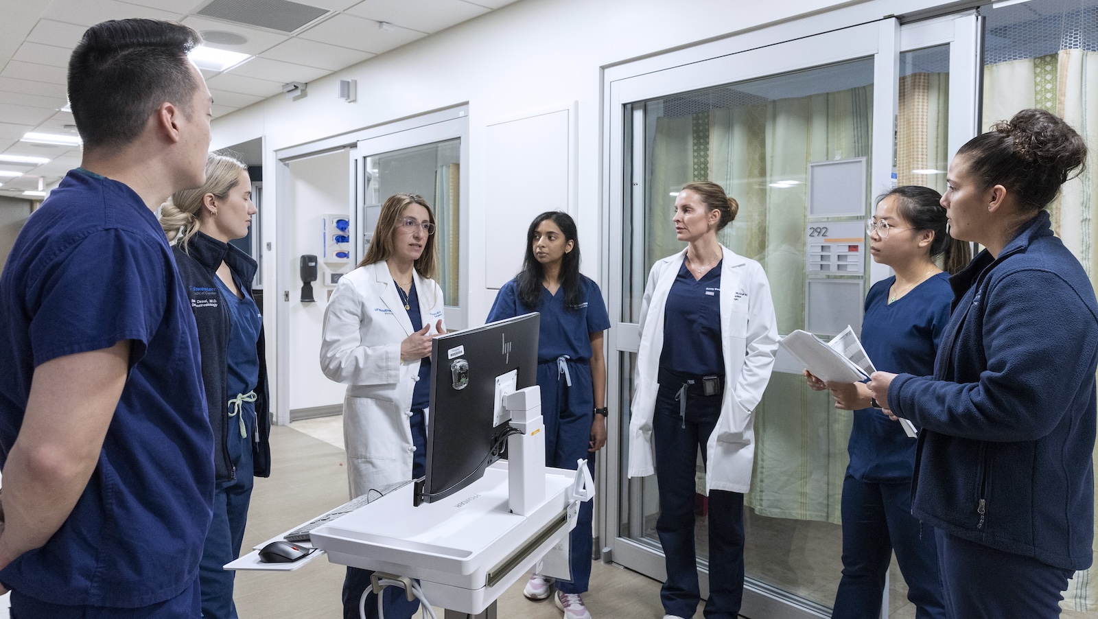 A group of medical professionals in blue scrubs and white coats gather for a discussion in a hospital hallway