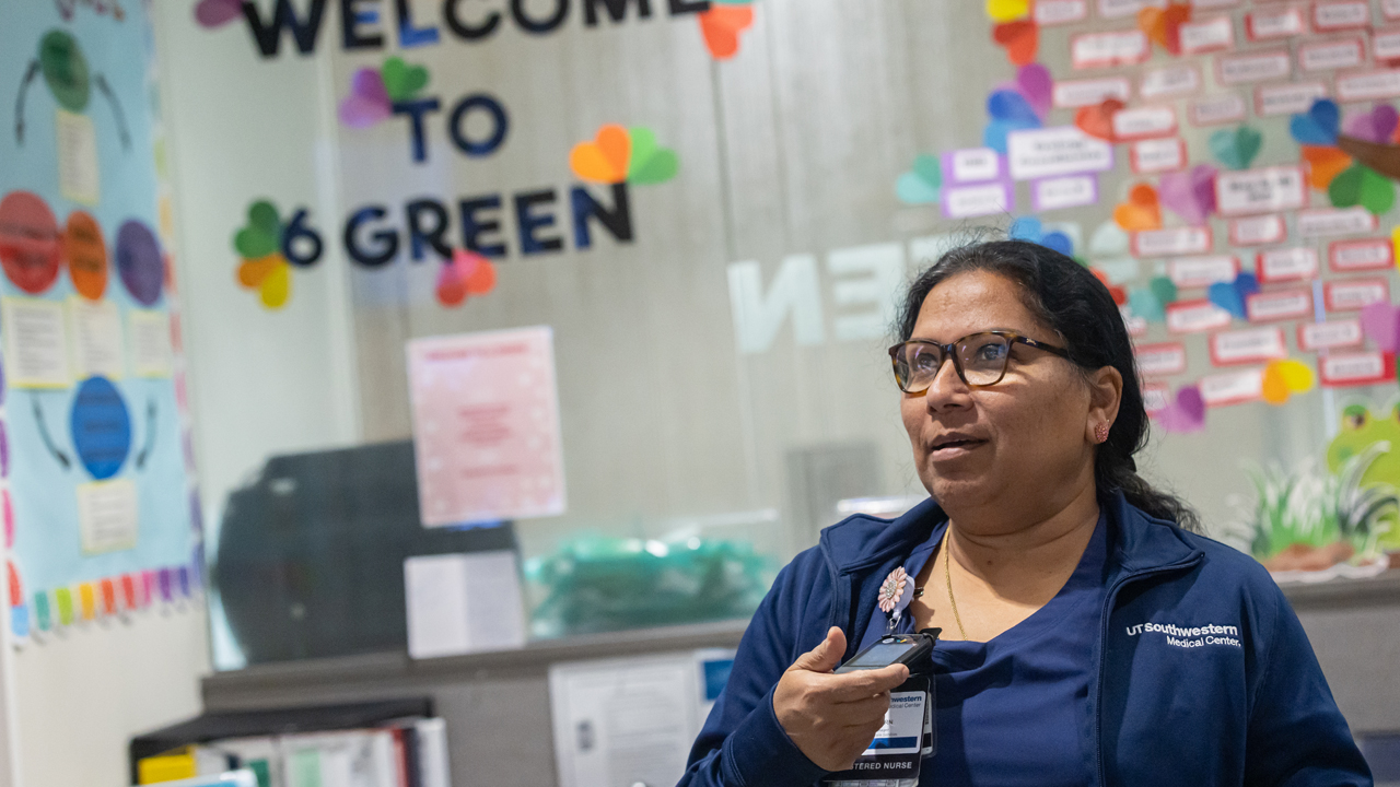 A female nurse in blue scrubs and a blue jacket standing in front of a wall covered in sticky notes.