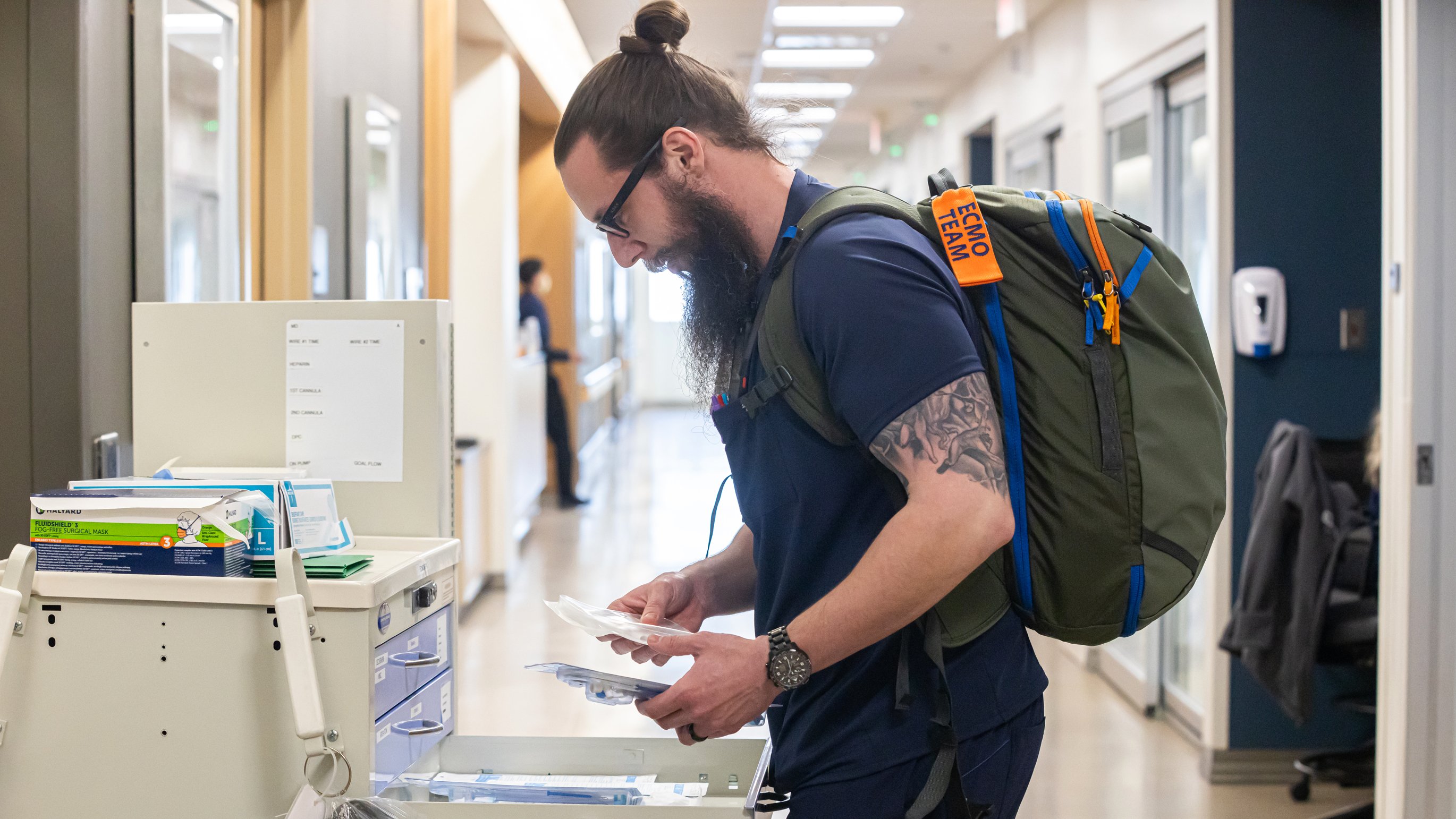A male nurse with glasses and a backpack gathering supplies from a drawer.
