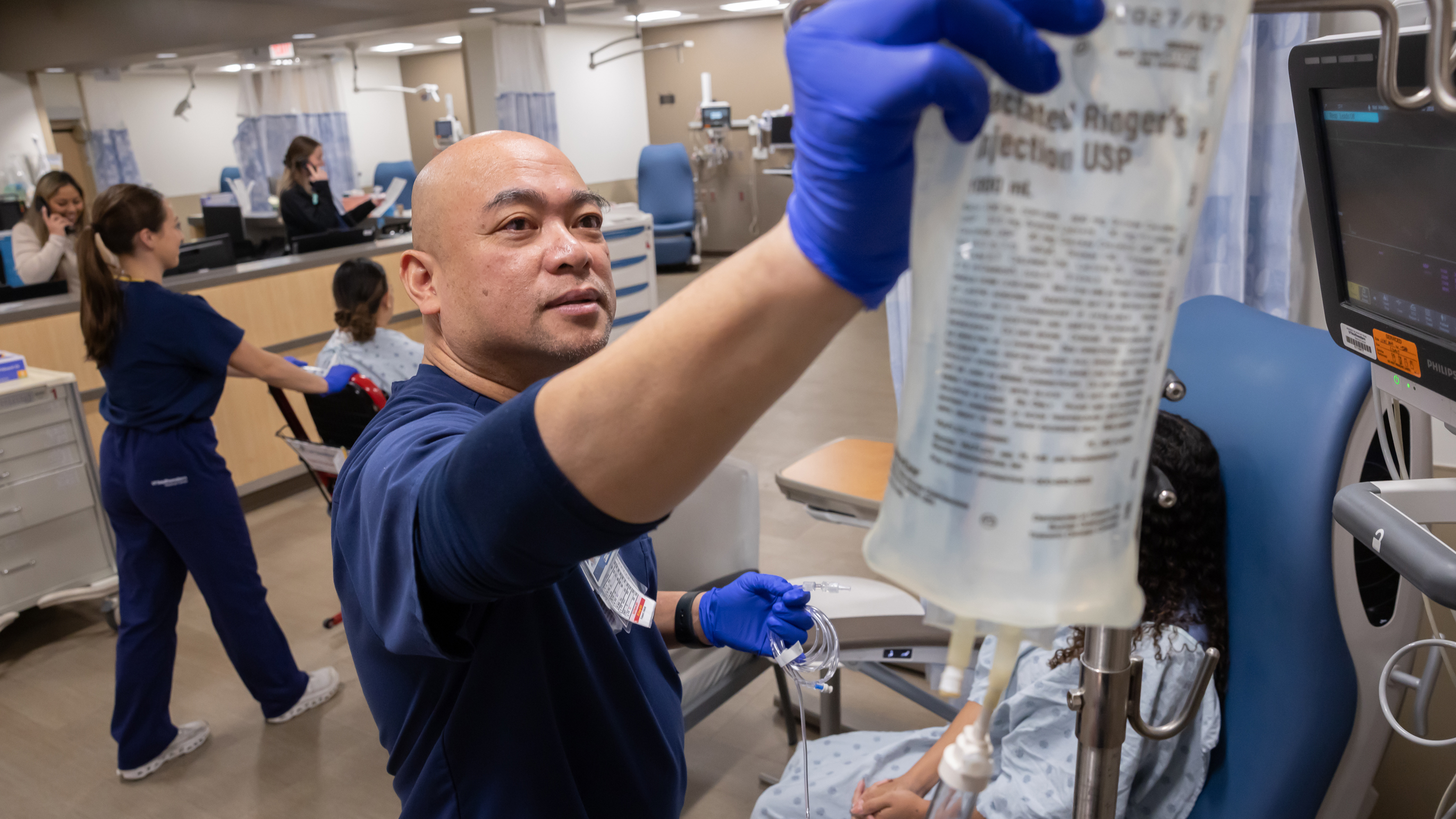 A male nurse in blue scrubs and blue gloves holds an IV bag up. In the background, another nurse is pushing a patient in a wheelchair and two other nurses are behind a desk.