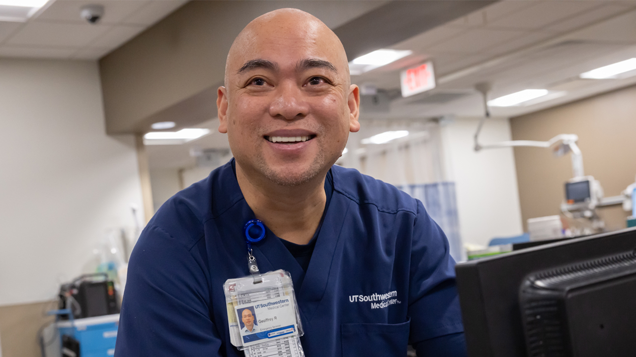 A male nurse standing at a computer, smiling at someone behind the camera.