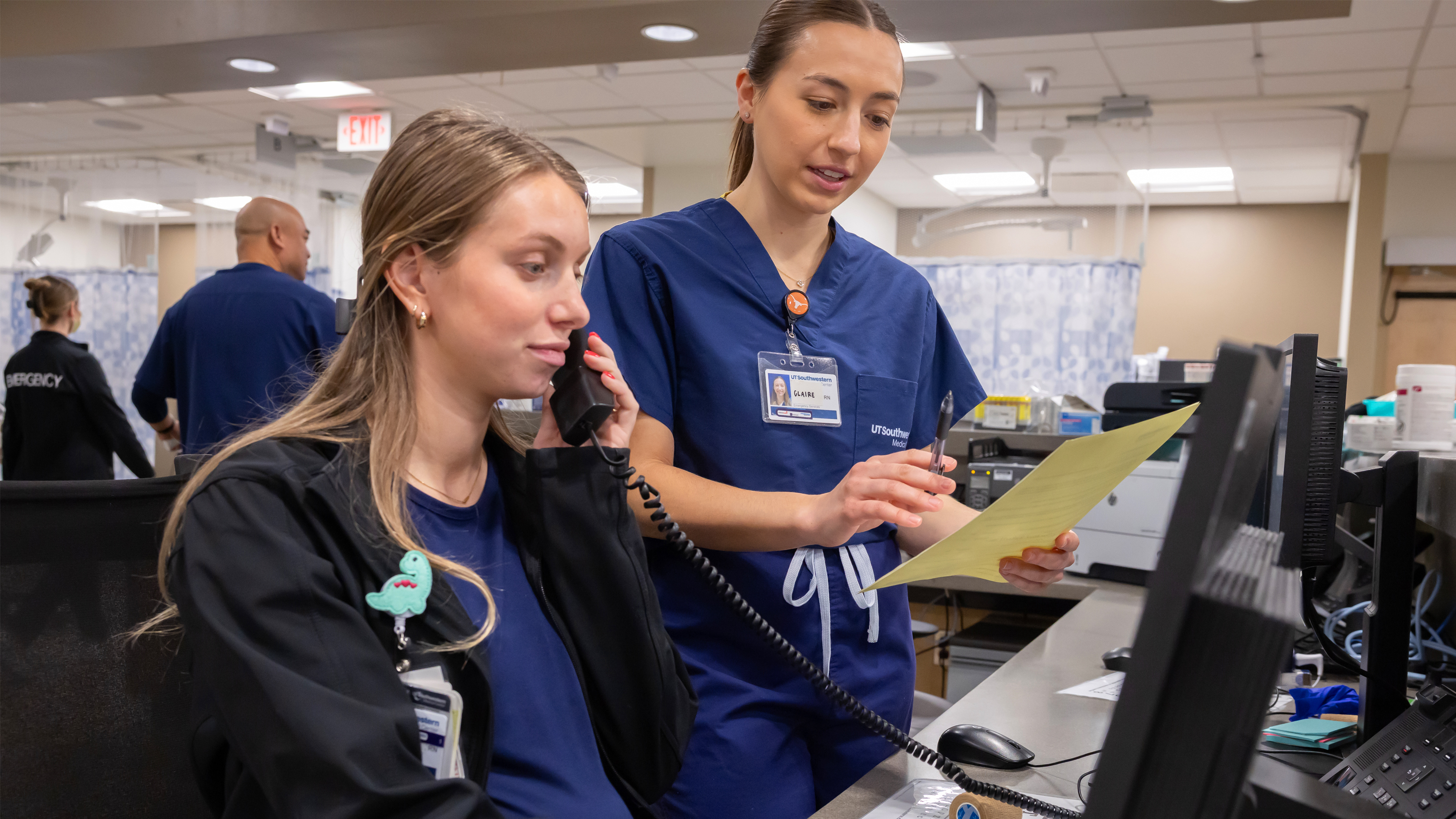 Two nurses next to each other at a nurse's station; one is on the phone while the other points to information on a sheet of paper