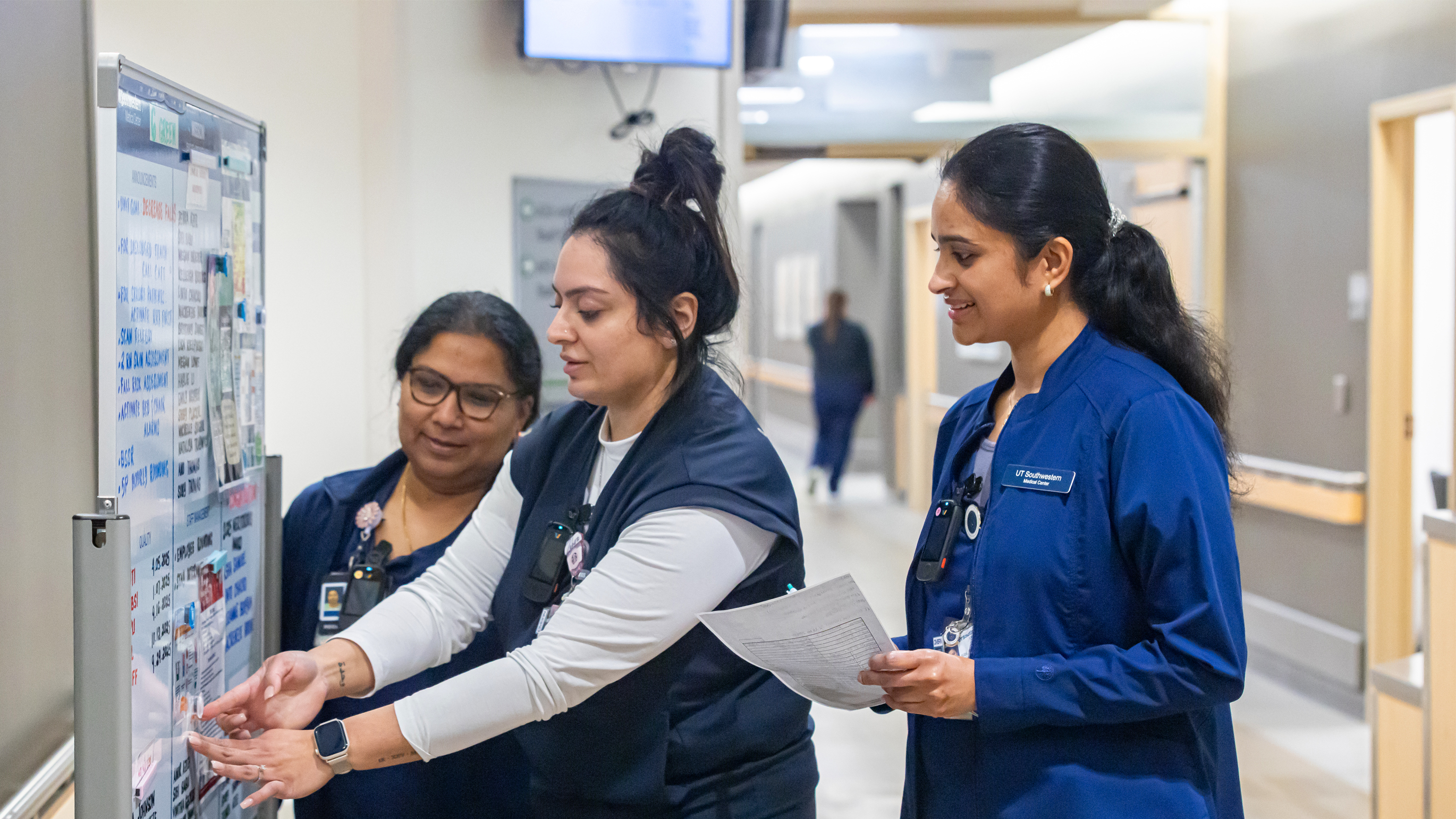 Three nurses standing in a hallway discussing information written on a white board.