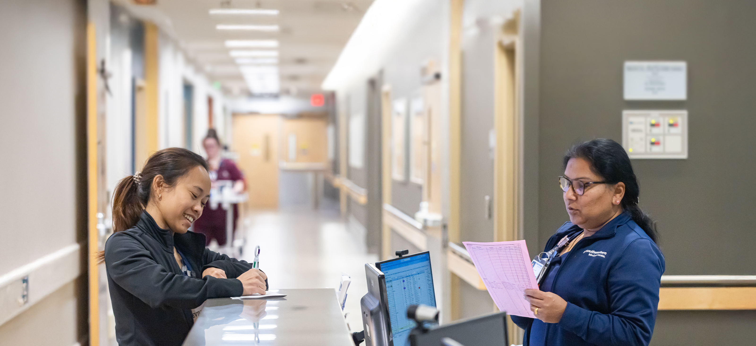 Two nurses standing on opposite sides of a desk. One is writing with a pen, and one is reviewing information on a pink sheet of paper.