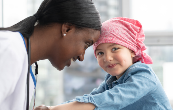 Female doctor gently touching foreheads with a young girl wearing a pink headscarf, both smiling warmly.