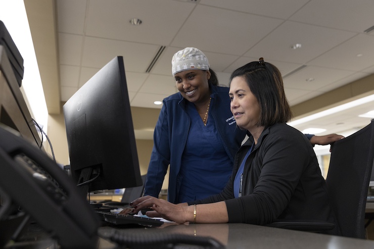 Two healthcare workers, one standing and one sitting, collaborate at a computer workstation in a hospital