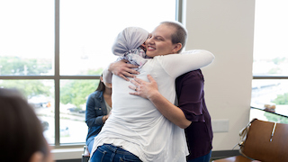 Two women embrace warmly in a bright room, one wearing a headscarf and the other smiling with her eyes closed