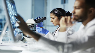 Two scientists work in a lab, with one examining a slide under a microscope and the other analyzing data on a computer screen