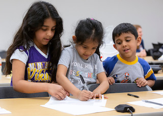 Three young children work together on a drawing at a table, with one child holding a pen and the others watching closely