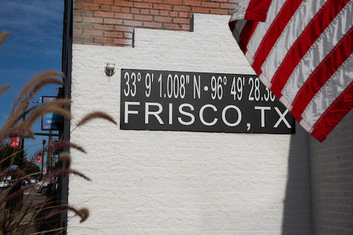 A white wall with a black sign displaying the coordinates and the name "Frisco, TX," accompanied by an American flag.