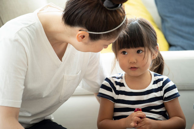 A mother leans in to speak gently to her young daughter, who looks thoughtful while sitting on a couch