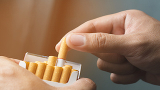 A hand pulls a cigarette from an open pack, with the rest of the cigarettes still inside the package