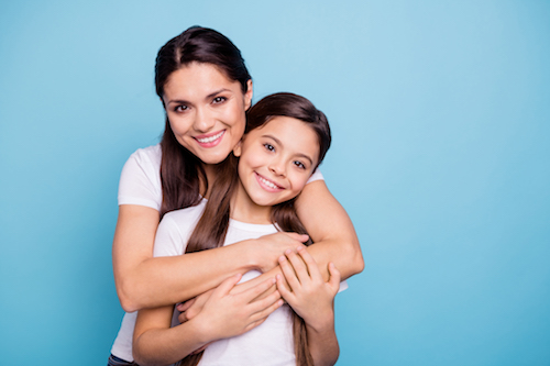 A mother and her daughter, both smiling, are hugging each other against a blue background.