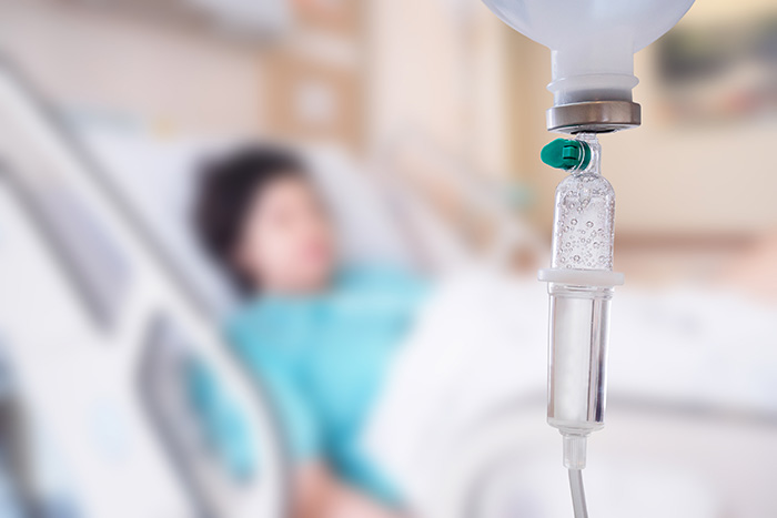 A close-up of an IV drip with a patient lying in a hospital bed in the blurred background