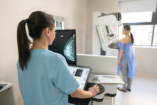 A healthcare professional monitors the results on a screen while a patient undergoes a mammogram