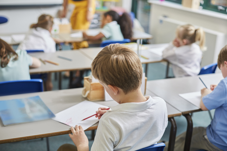 A classroom of young students focuses on their work, with a boy in the foreground writing in a notebook