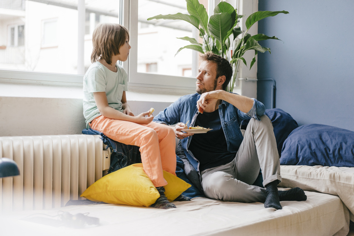 A father and his young child sit on a couch in a bright living room, engaged in a meaningful conversation