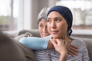 A woman wearing a headscarf is comforted by an older woman who embraces her from behind while they sit together on a couch