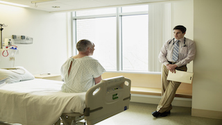 A doctor stands by the window in a hospital room, speaking with a patient who is sitting on the edge of a bed