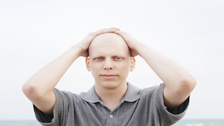 A man with a shaved head stands outdoors, looking directly at the camera while holding his hands on top of his head