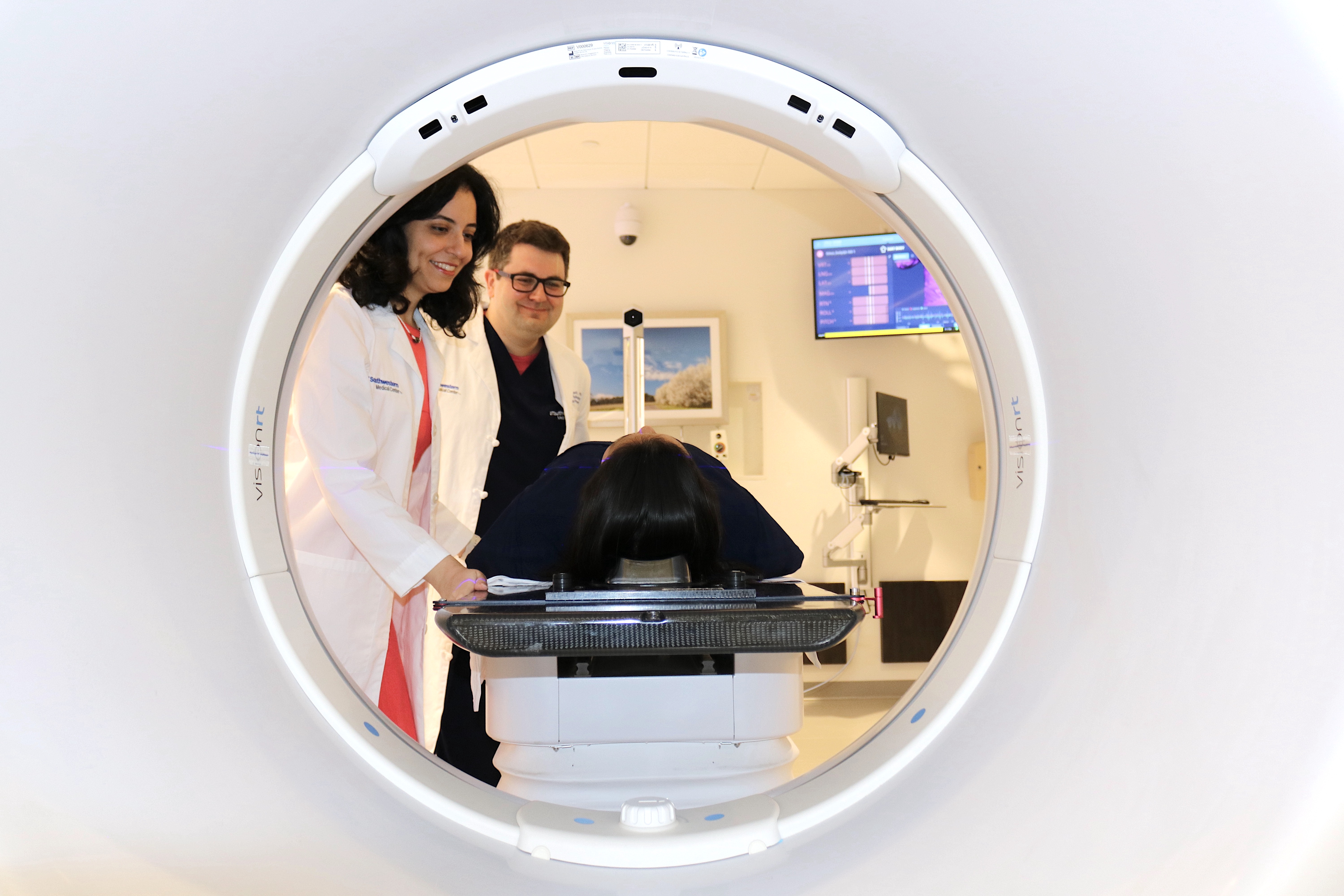 Two healthcare professionals are positioning a patient inside a medical imaging machine, viewed from the perspective of looking through the machine's circular opening.