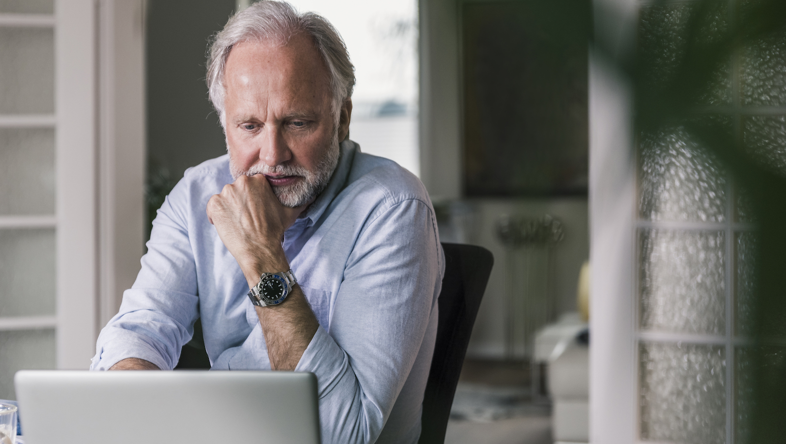 A man with a thoughtful expression is working on a laptop in a home or office setting.