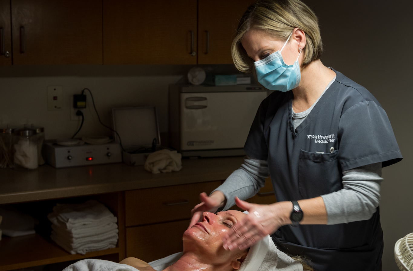 A skincare professional is giving a facial massage to a client lying down on a treatment bed, with towels and skincare products in the background.