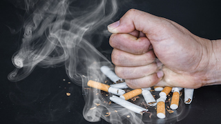 A clenched fist crushes a pile of broken cigarettes, with smoke still rising from them