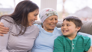 A woman, an elderly woman wearing a headscarf, and a young boy sit close together on a couch, smiling and embracing each other