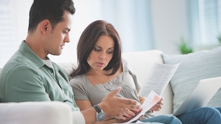 A couple sits together on a couch, closely reviewing a document with a serious and focused expression