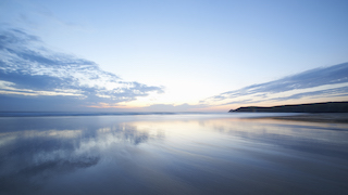 A serene seascape at dawn, with a calm sea reflecting a soft, pastel sky and wispy clouds.