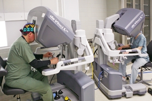 Two surgeons are seated at consoles operating a da Vinci robotic surgical system in a clinical setting.