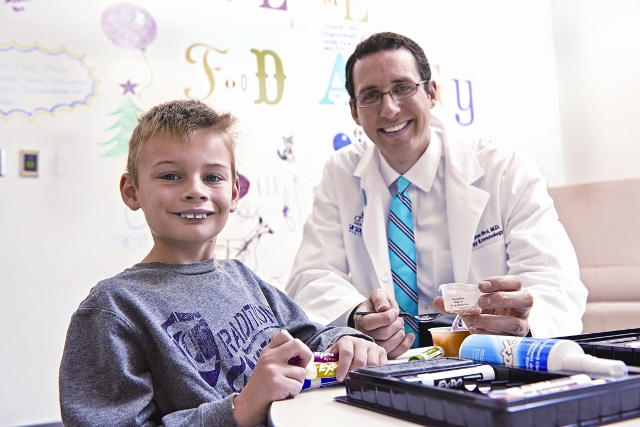 A doctor is smiling and sitting next to a young boy in a clinical setting, holding medical instruments.
