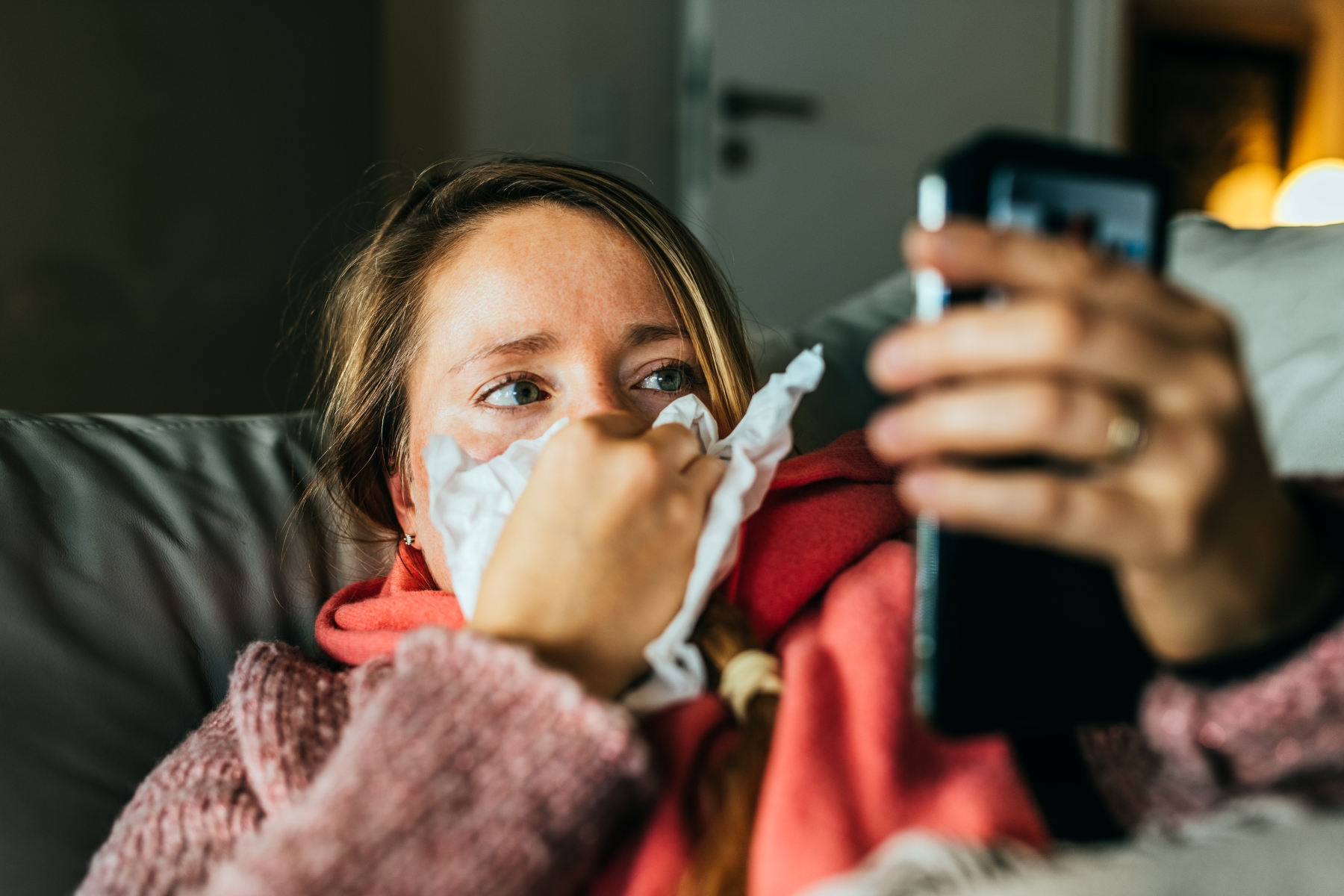 Sick woman holding tissue to nose with one hand and checking smartphone in the other hand