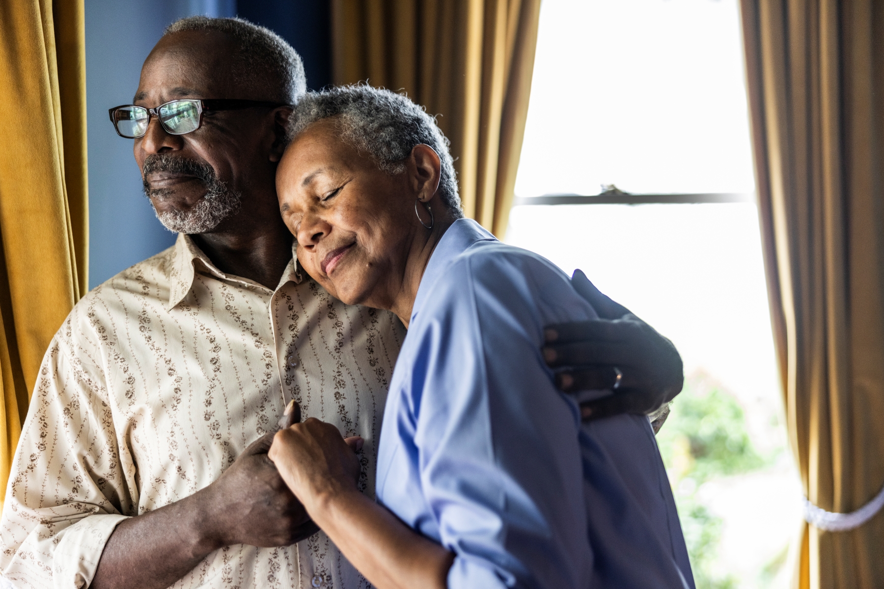 Older man and woman sharing an embrace in front of window with curtains open