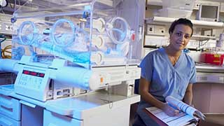 A healthcare professional in scrubs is seated beside an incubator with a premature baby inside, surrounded by medical equipment.