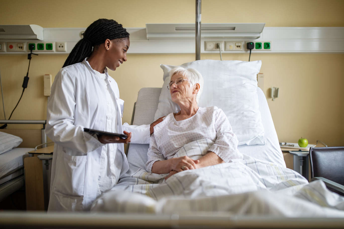 A healthcare professional in a white coat is speaking with an elderly patient who is lying in a hospital bed