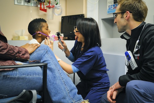A healthcare professional is examining a child's mouth with a light, while another adult observes.