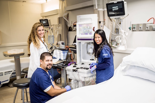 A clinical setting with three medical professionals smiling, preparing a medical device in a treatment room.