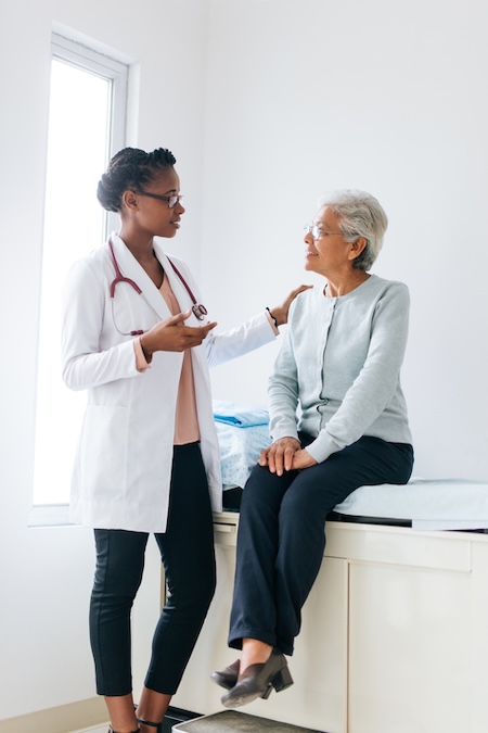 A senior female patient sitting on a consulting bed and listening to a young female doctor standing next to her.