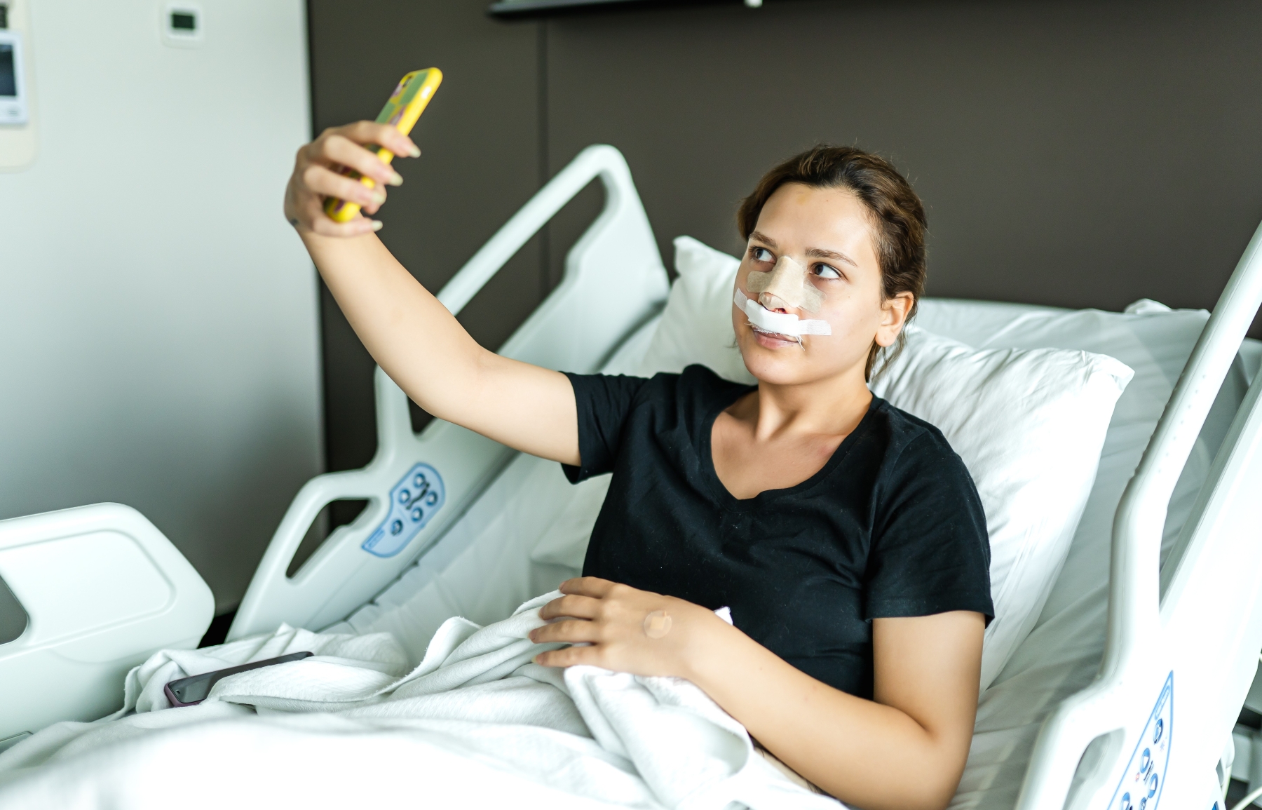 A woman with tape on nose and upper lip taking selfie while sitting in hospital bed after surgery