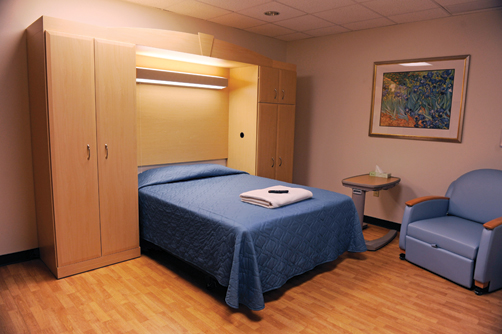 A sleep room featuring a bed with blue bedding, a comfortable armchair, and a calm, organized setting designed for conducting sleep studies related to sleeping and breathing disorders.