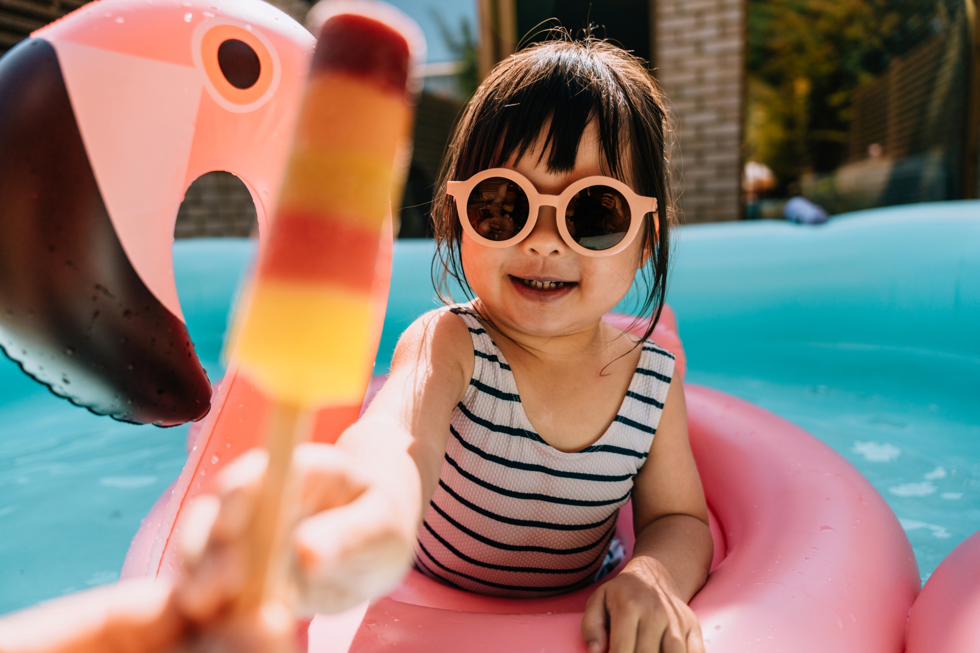 Young girl wearing sunglasses in pool with floatie and being offered a popsicle