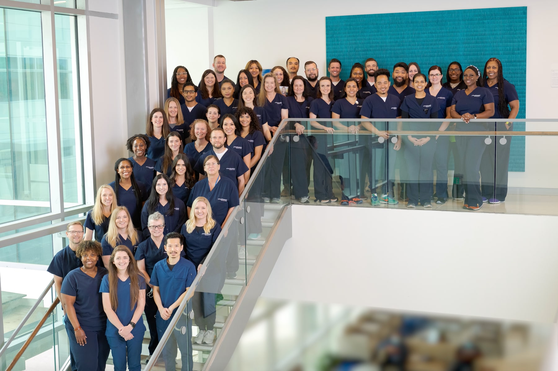 A large group of radiation therapists, all wearing matching navy blue shirts, are standing on a staircase inside a modern building, posing for a group photo.