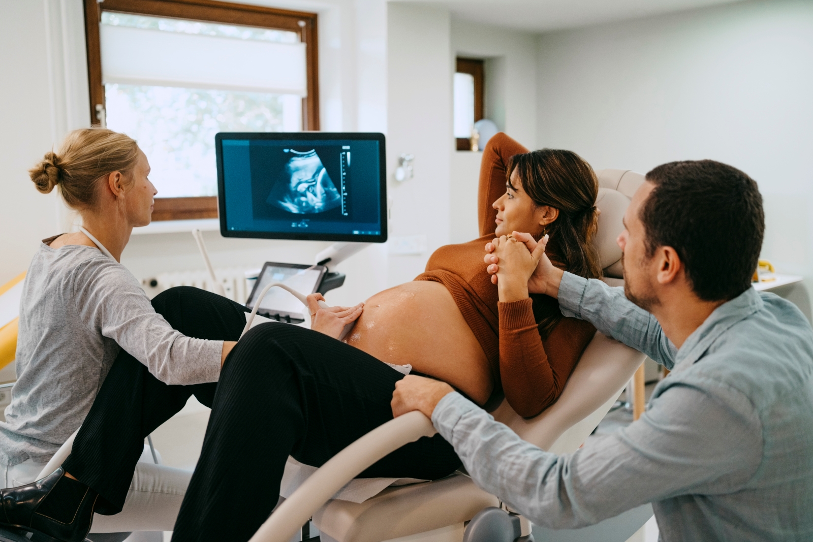 clinician performing ultrasound on woman who is pregnant. Patient is holding hands with a man. All are looking at monitor.