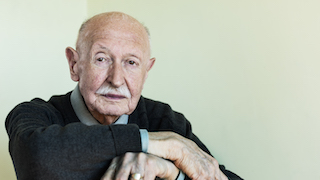 An elderly man sitting pensively with hands crossed.