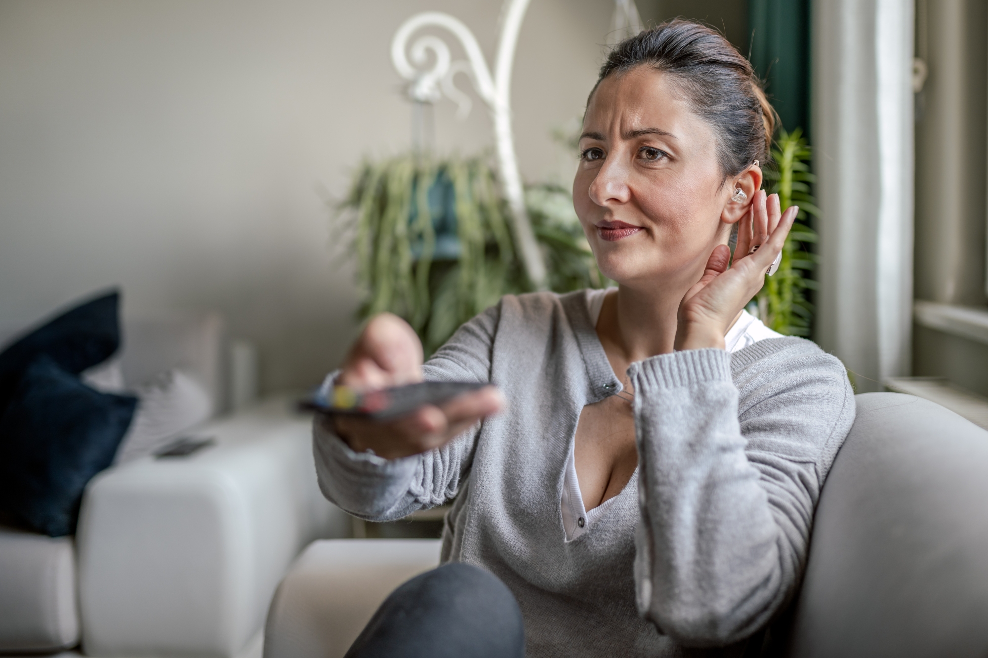 Woman on couch cupping ear with hand while pointing remote to a TV