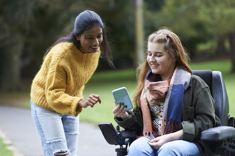 Two young women, one in a wheelchair, are outdoors looking at a phone together.