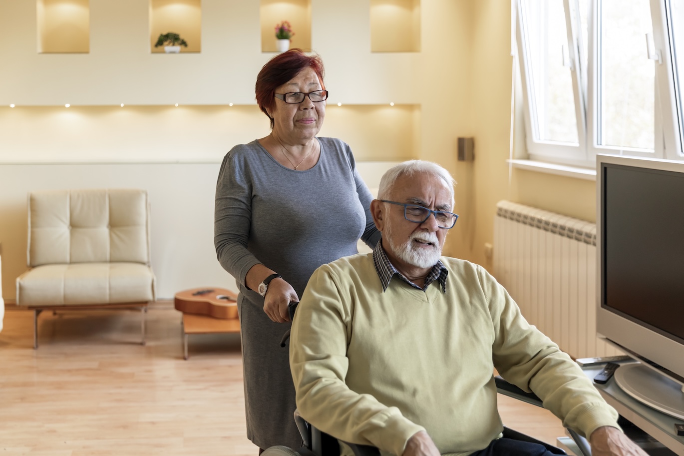 Wife pushing her husband in a wheelchair in their home.